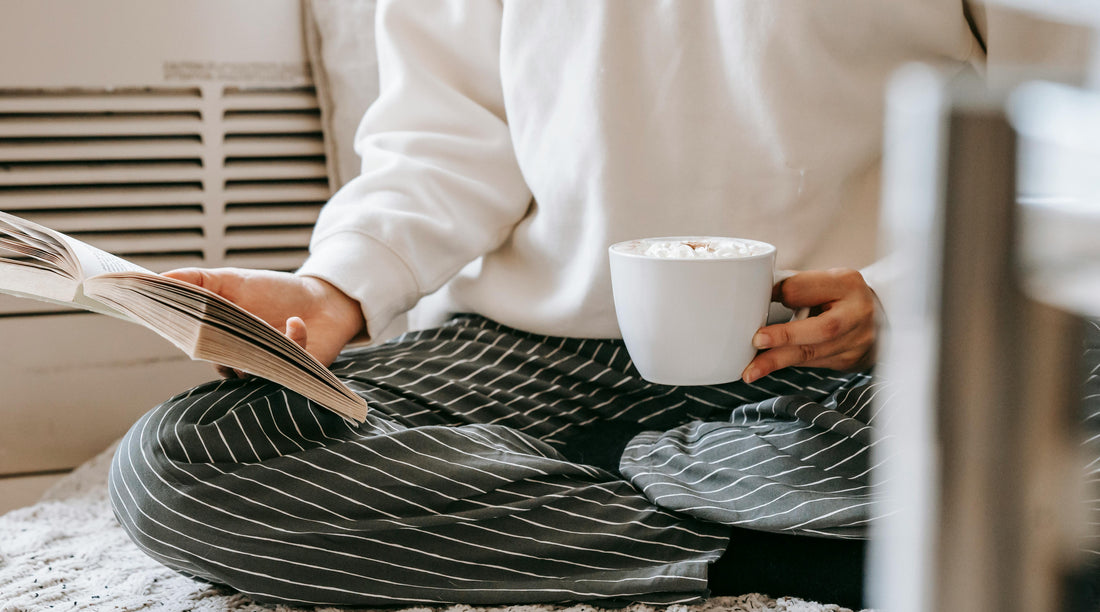 A woman in a cream sweatshirt and striped pants sits cross-legged while reading an open book and holding a warm cup of coffee with whipped cream, photographed in soft natural light.