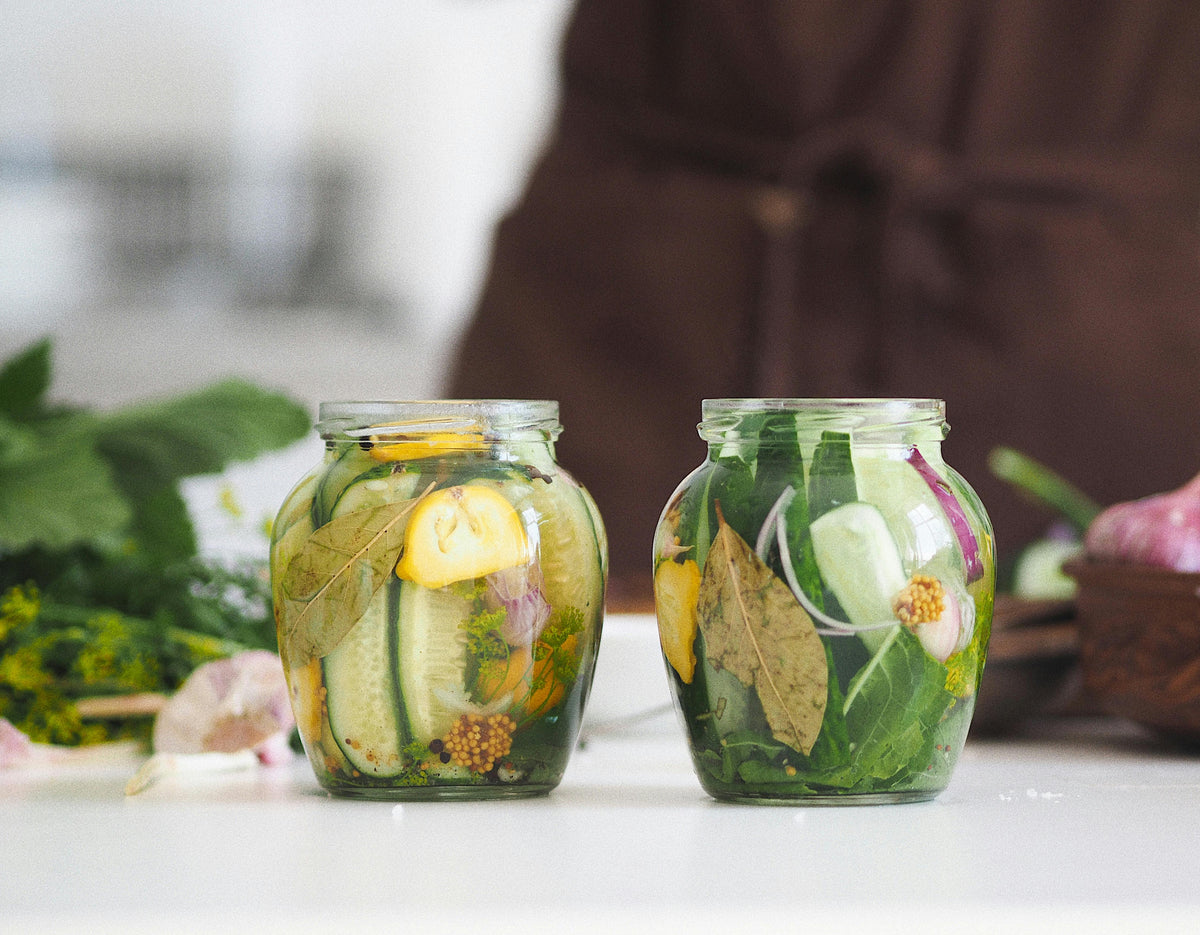 Two glass jars of fermenting cucumbers with bay leaves, dill, lemon, garlic, and mustard seeds on a cream kitchen counter in soft natural light, with fresh herbs and a person preparing food softly blurred in the background.